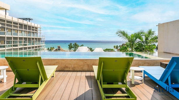 A rooftop pool area with two green lounge chairs and a blue one, overlooking a clear blue ocean and a resort building. - Sabor A Miel by Stella Rentals (Playa del Carmen)