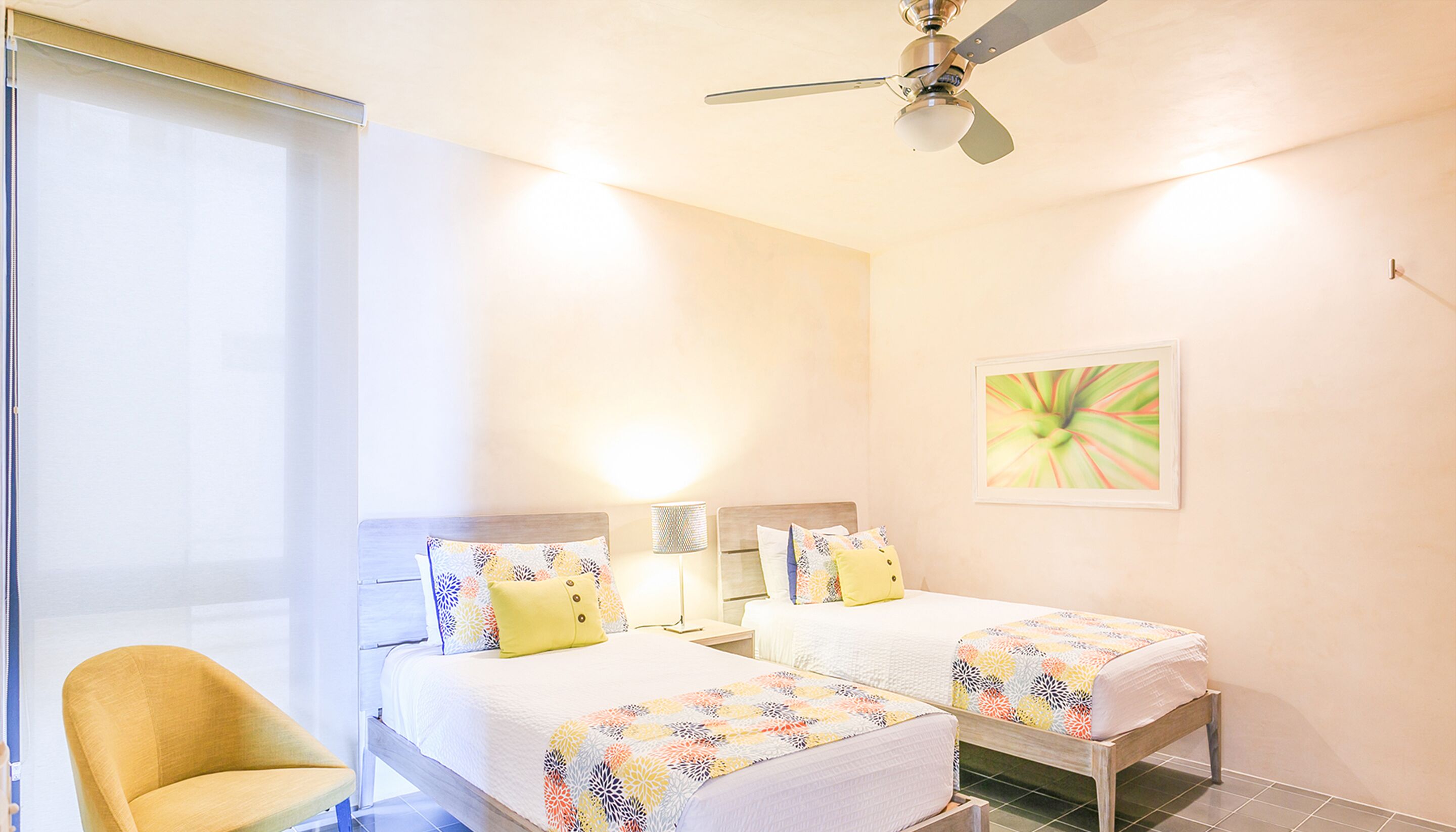 Une chambre d’hôtel avec deux lits, un ventilateur de plafond, une chaise, une table de chevet et une œuvre d’art murale.
