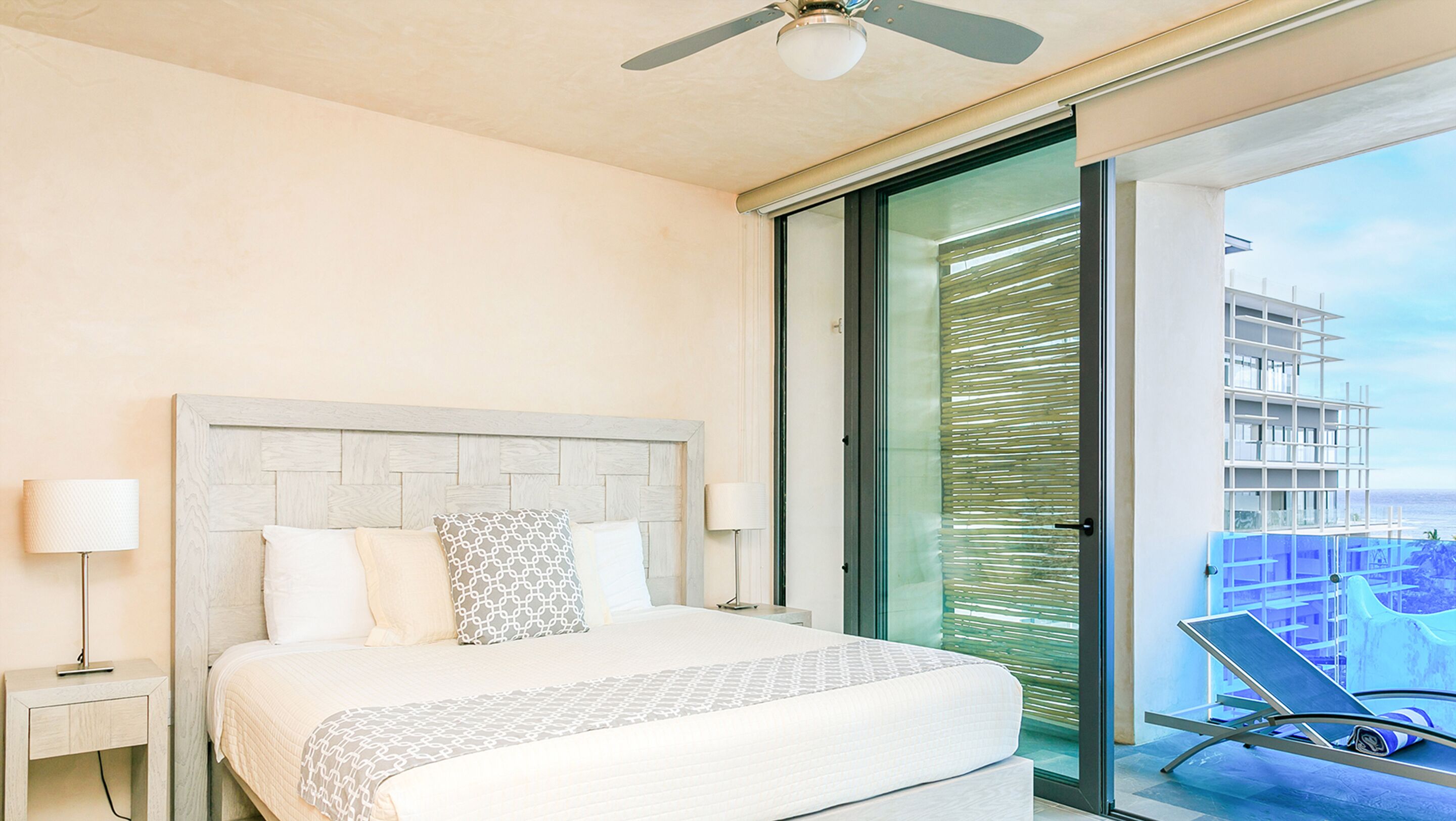 Une chambre d’hôtel comprenant un lit, une table de chevet, un ventilateur de plafond, une grande fenêtre donnant sur un immeuble et un balcon avec une chaise.