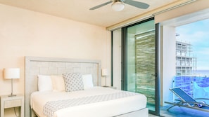Une chambre d’hôtel comprenant un lit, une table de chevet, un ventilateur de plafond, une grande fenêtre donnant sur un immeuble et un balcon avec une chaise.