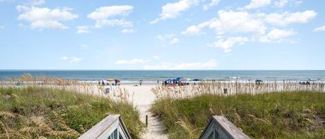 A beach path leading to the ocean with beachgoers and umbrellas.