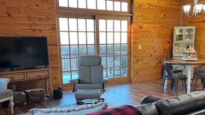 A cozy cabin interior with a large window, a television, a dining area, and a sofa with a plaid blanket.