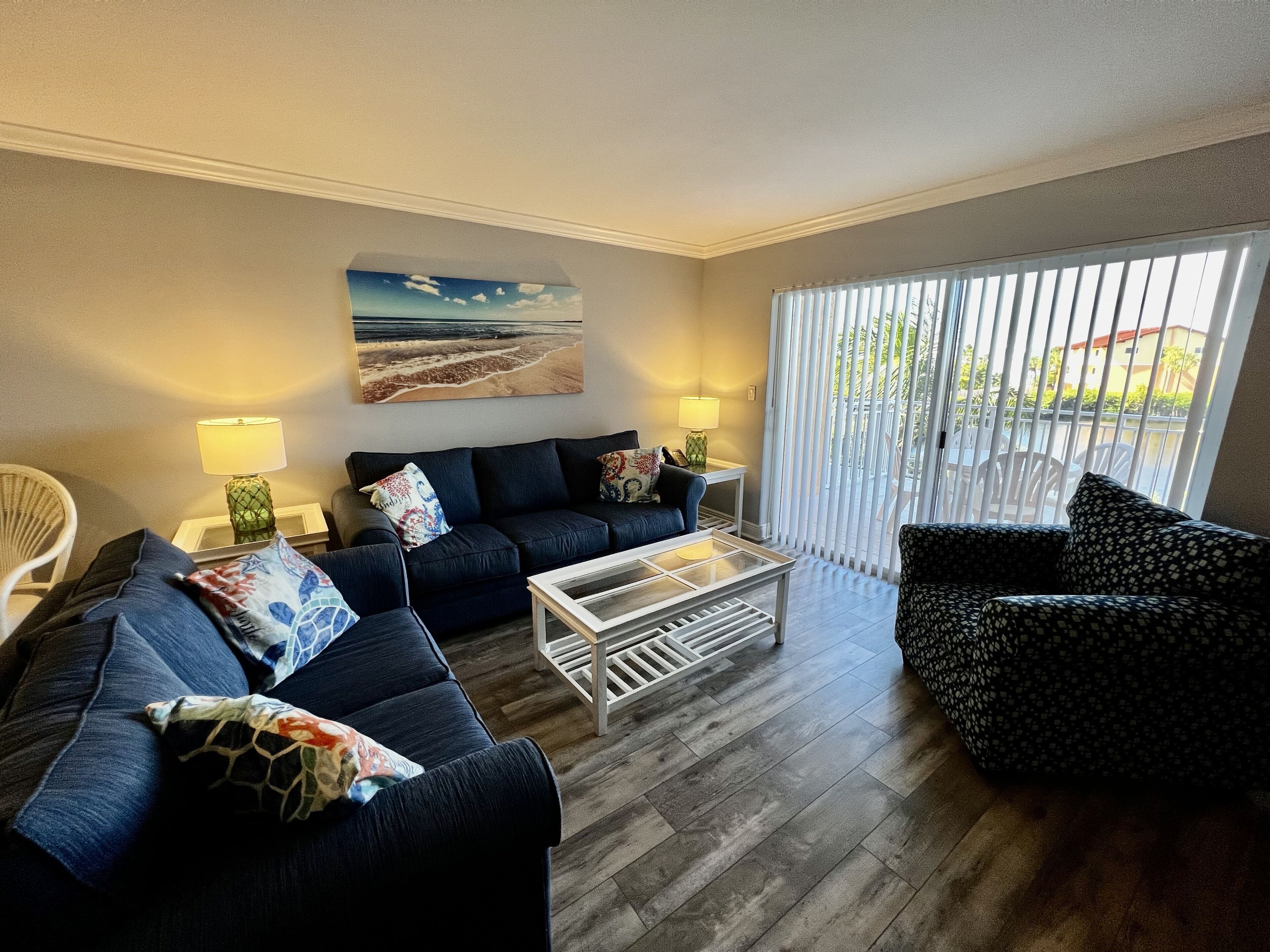 Een woonkamer met een donkerblauwe bank, een fauteuil met een patroon, een glazen salontafel en een schilderij van een strandgezicht aan de muur.