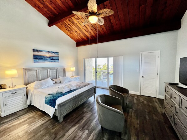 A bedroom with a wooden ceiling, a bed with white linens, a chair, a TV, and a view through the door.