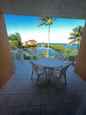 A round table with chairs on a balcony overlooking a body of water with palm trees and a building in the background. - Spacious condo, great location in paradise! (Key Largo)