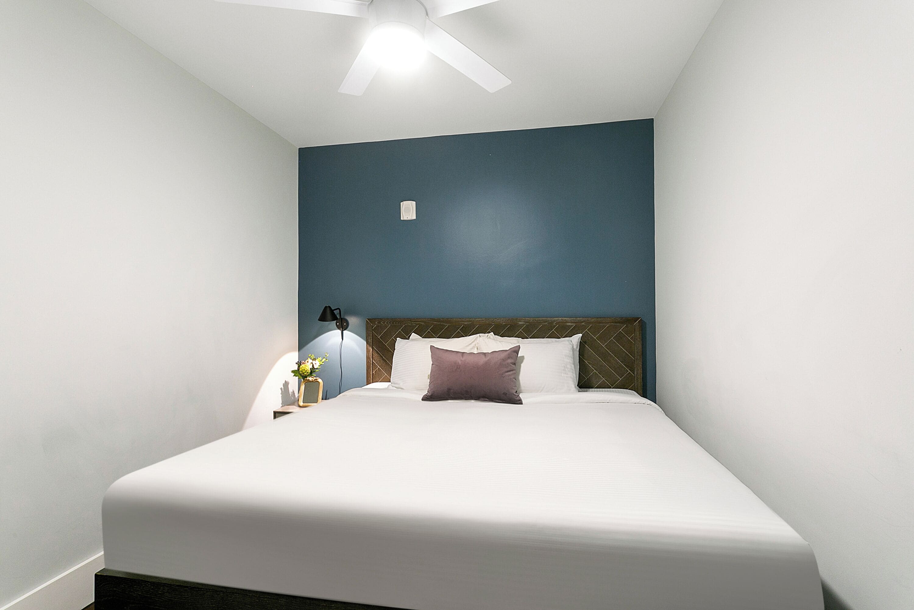 A neatly made bed with white bedding and a single purple pillow, a bedside lamp, and a small nightstand with a vase of flowers.