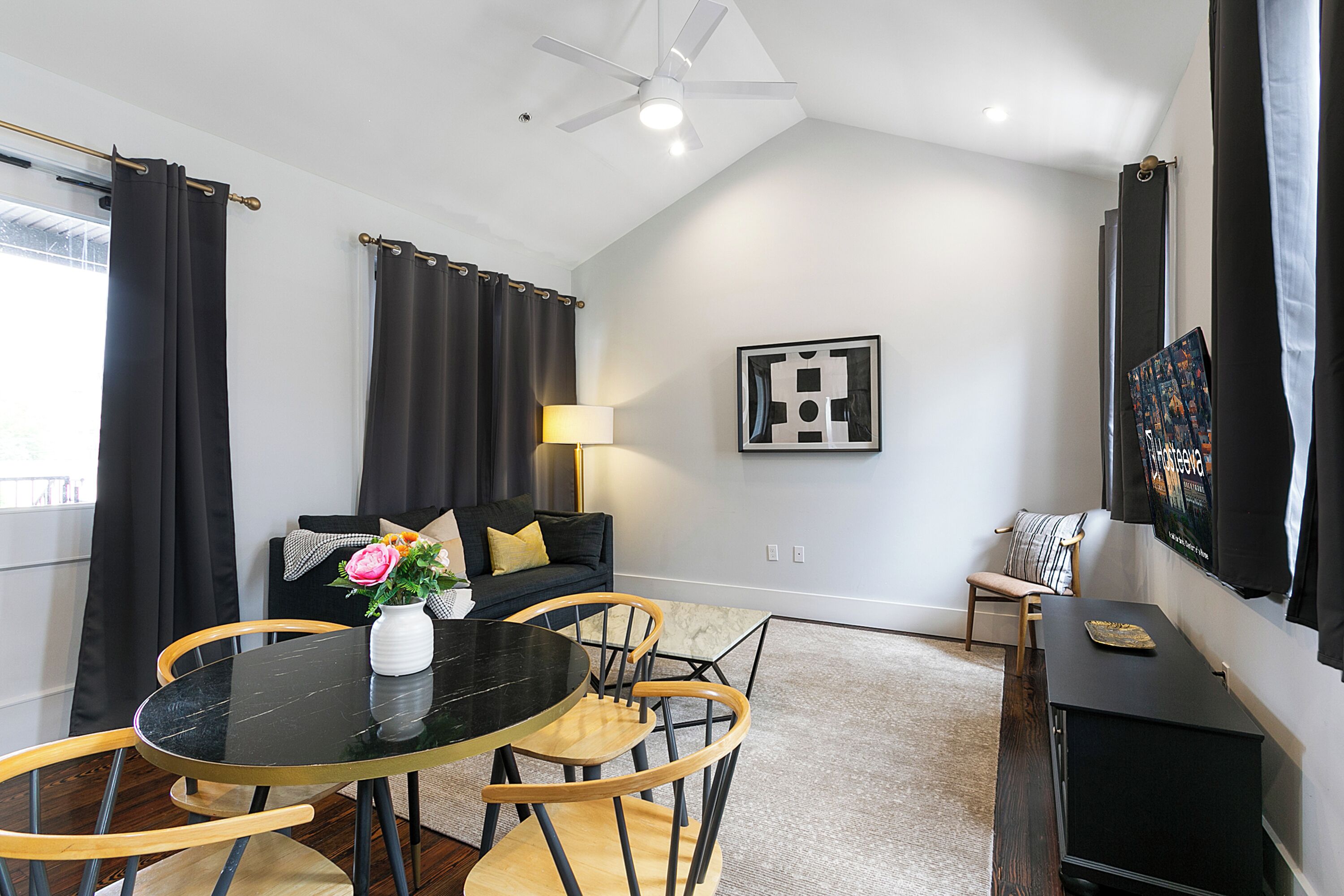 Un salon moderne avec une table ronde noire, des chaises en bois, un canapé noir avec des coussins, un lampadaire et un tableau encadré au mur.