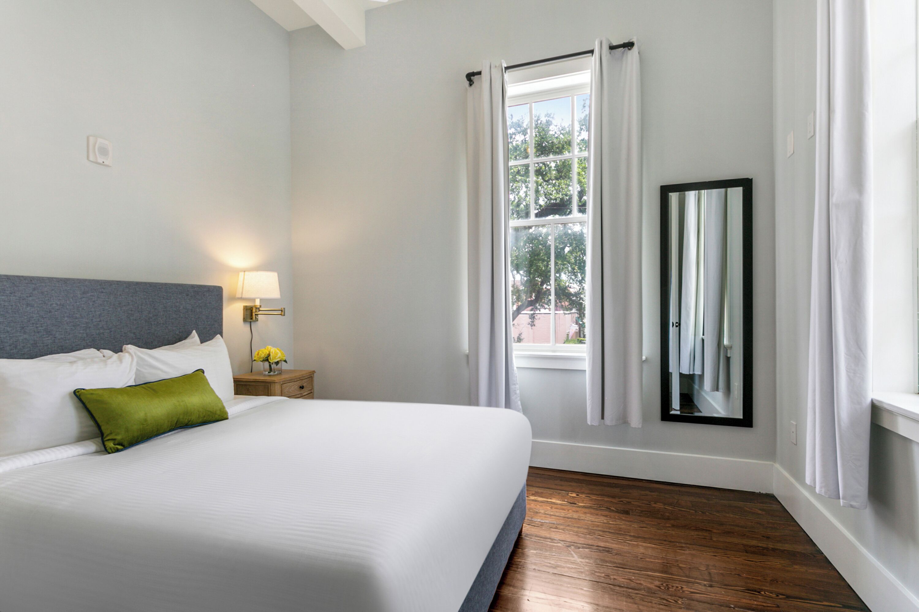 A hotel room with a bed, a nightstand with a lamp, a window with curtains, and a mirror on the wall.