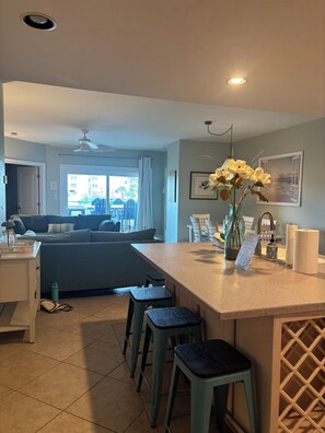 A modern kitchen with a central island, bar stools, a dining area with a sofa, and a view of the outdoors.
