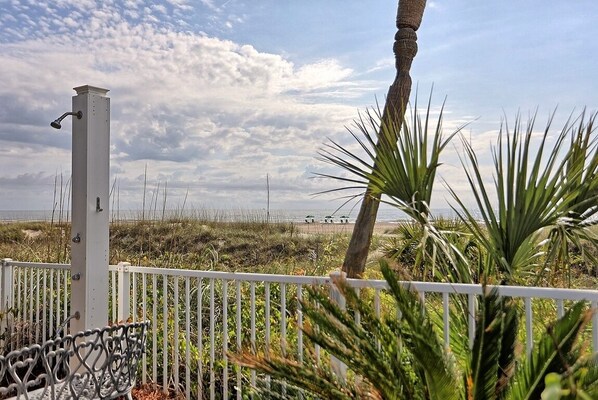 A white metal fence with a metal bench and a tall white structure, possibly a light post, in a coastal area with dunes and a beach in the background.