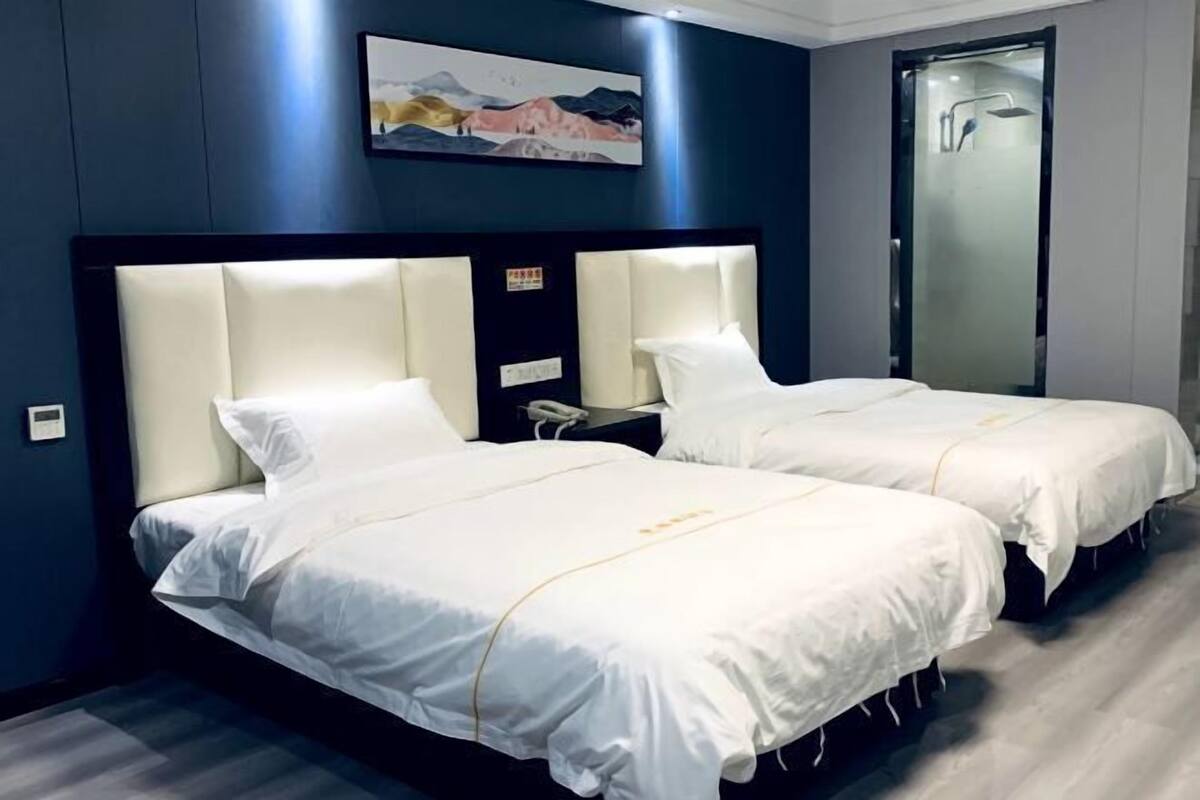 Hotellihuone, jossa on kaksi sänkyä, suuri päätyseinä, taulu seinällä ja lasinen suihkuovi.