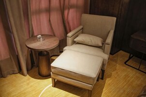 Una habitación pequeña y acogedora con un sillón individual color beige y un reposapiés, una mesa redonda de madera, y un telón de fondo.