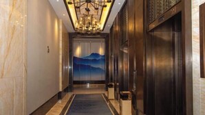 A modern hotel lobby with a marble floor, a blue patterned carpet, a large chandelier, and an elevator with a mountain mural.