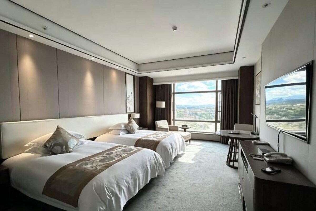 Une chambre d’hôtel avec deux lits, un bureau, une chaise et une grande fenêtre donnant sur la ville.