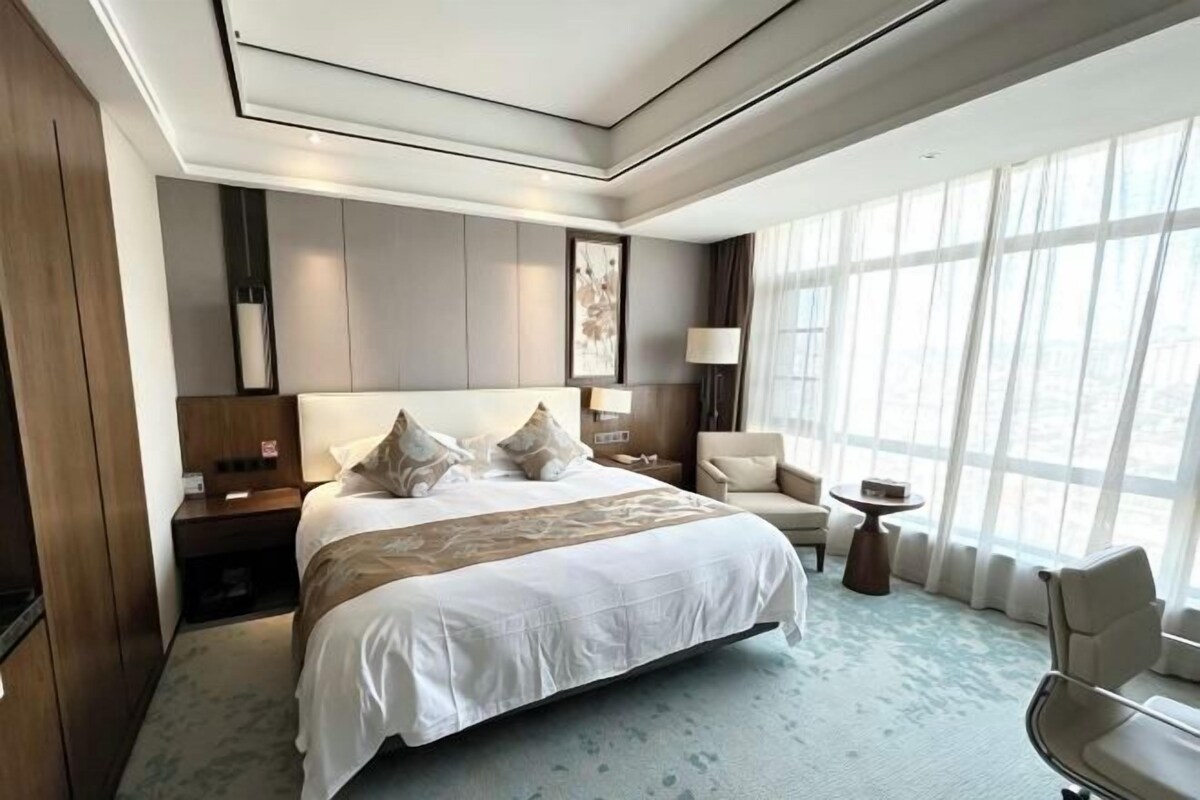 Une chambre d’hôtel avec un grand lit, des tables de chevet, une chaise, une petite table et une fenêtre avec des rideaux.