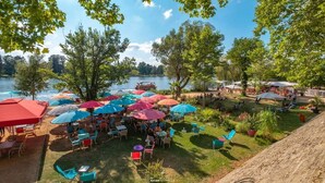 An outdoor seating area with umbrellas and tables by a lake, surrounded by trees.
