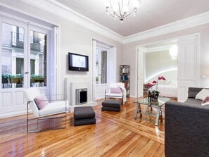 Een ruime woonkamer met een open haard, een aan de muur bevestigde televisie en een groot raam met uitzicht op de straat.