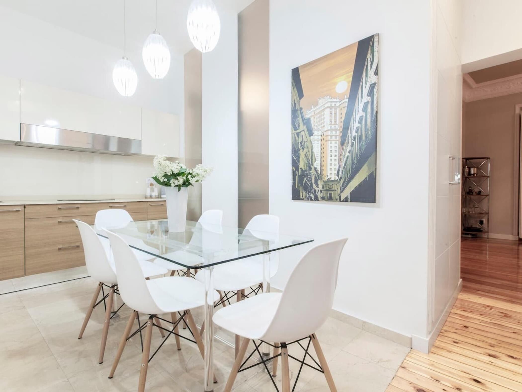 Une cuisine moderne avec des meubles blancs, une table à manger en verre et des chaises blanches. On peut voir un vase avec des fleurs sur la table et un tableau au mur.