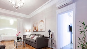 Ein modernes Wohnzimmer mit grauem Sofa, einem Couchtisch und einer Deckenleuchte. Ein Bett mit weißer Bettwäsche und eine Pflanze stehen daneben.