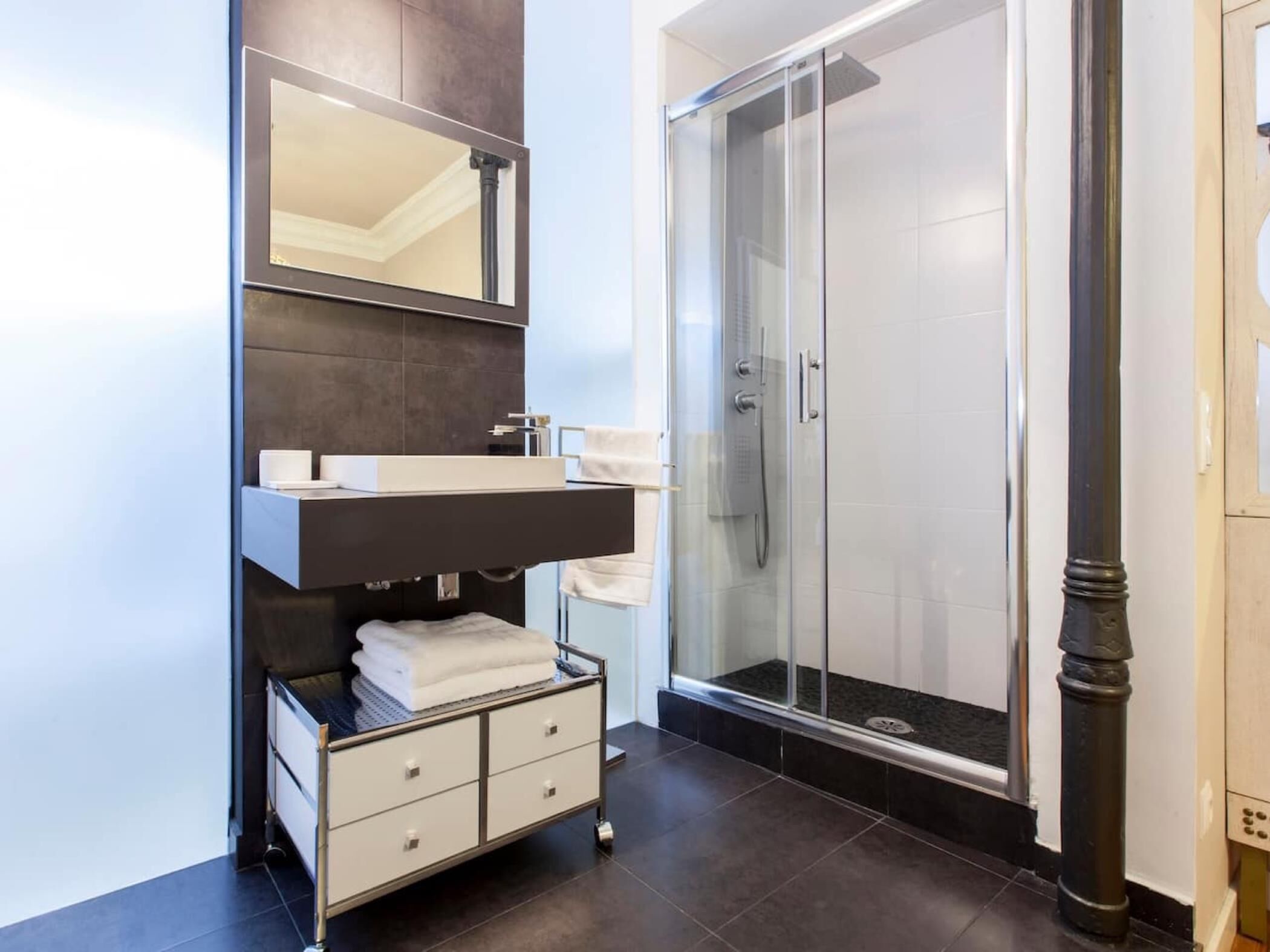 Une salle de bain moderne avec un carrelage foncé au sol, un lavabo blanc avec un miroir au-dessus, et une douche séparée par des parois en verre.
