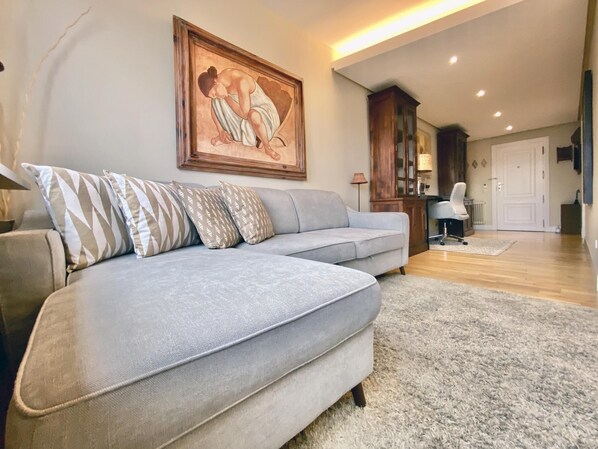 A living room with a grey sectional sofa, patterned pillows, a framed artwork above the sofa, a wooden cabinet, and a lamp.