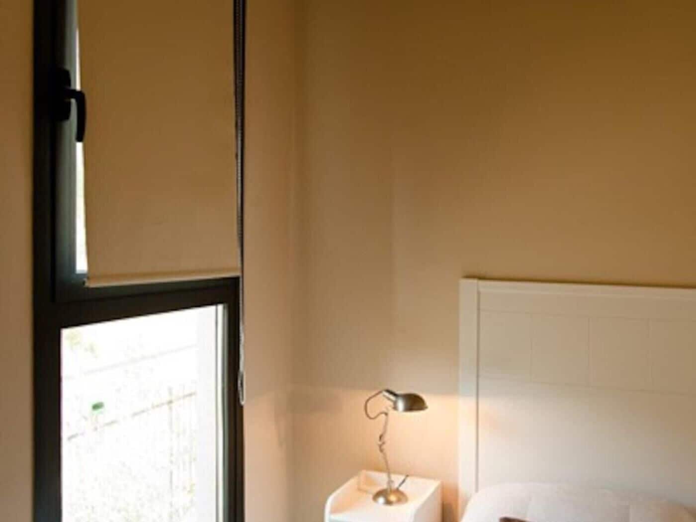 Habitación de hotel con cama, mesita de noche con lámpara y ventana con persianas.