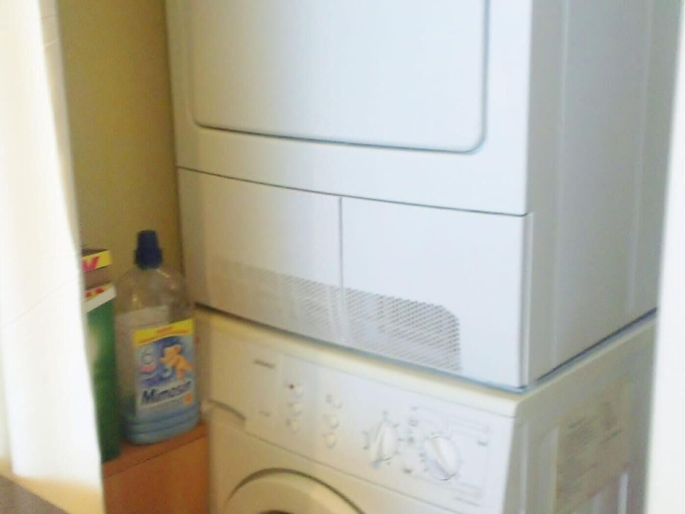Lavadora blanca compacta en un espacio de lavandería pequeño, con una botella de detergente y una caja cerca.