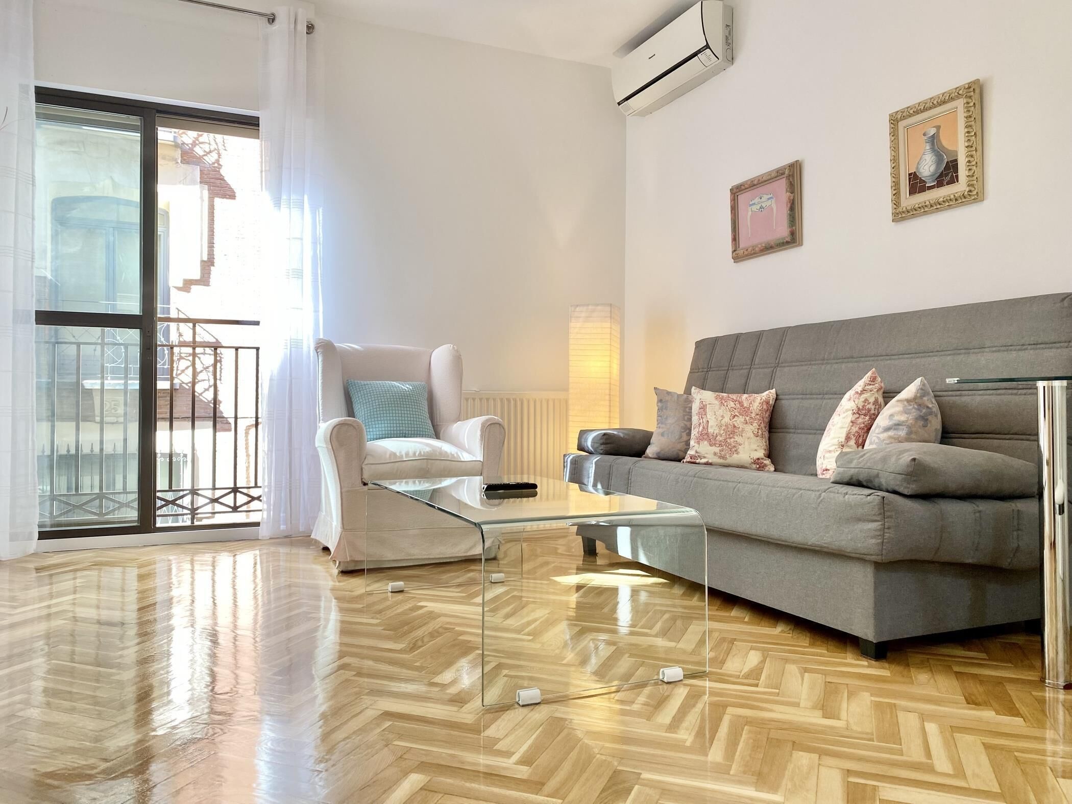 A modern living room with a grey sofa, a white armchair, and a glass coffee table. There is a balcony with a view of buildings outside.