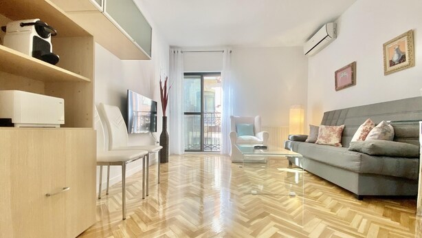 A modern living room with a herringbone floor, a grey sofa, a white armchair, and a glass coffee table. - Charming Gran Vía V - in the City Center (Madrid)