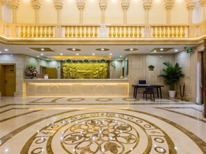 Un vestíbulo de hotel con una recepción, un diseño decorativo en el suelo y un balcón con barandillas doradas.