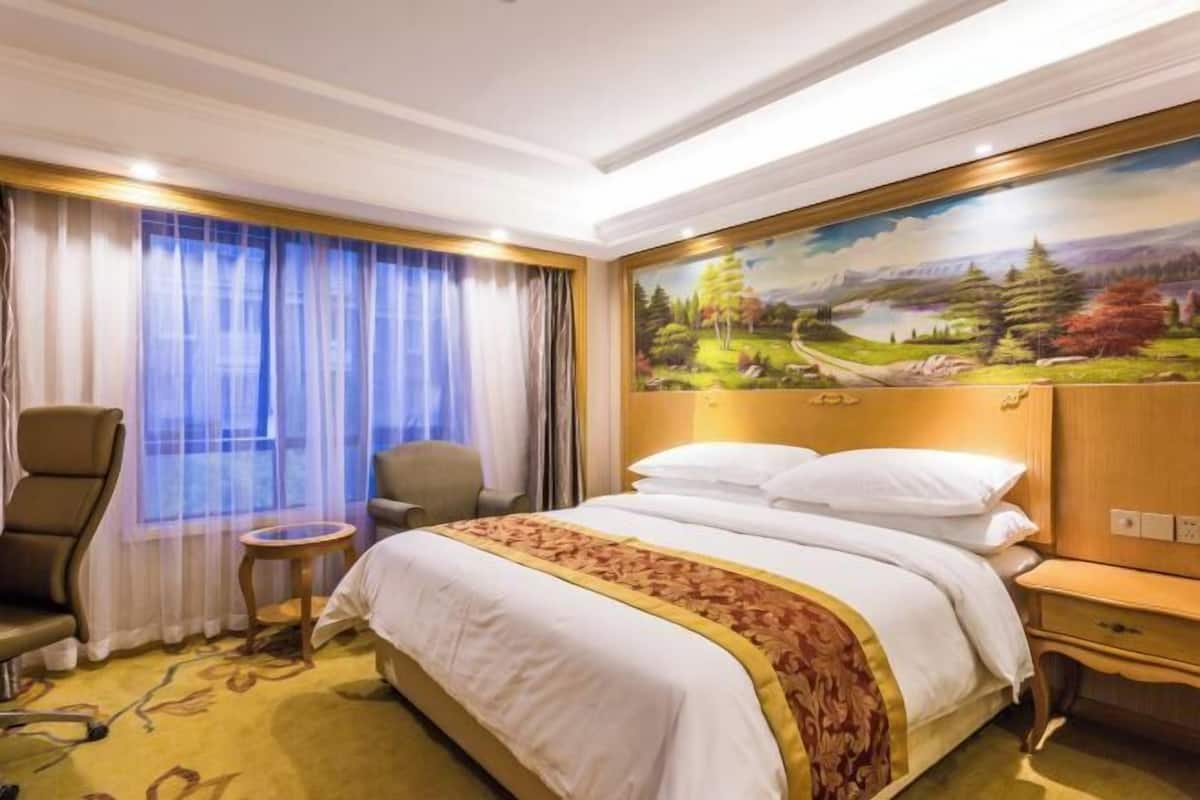 A hotel room with a large bed, a desk, a chair, and a painting of a landscape on the wall.