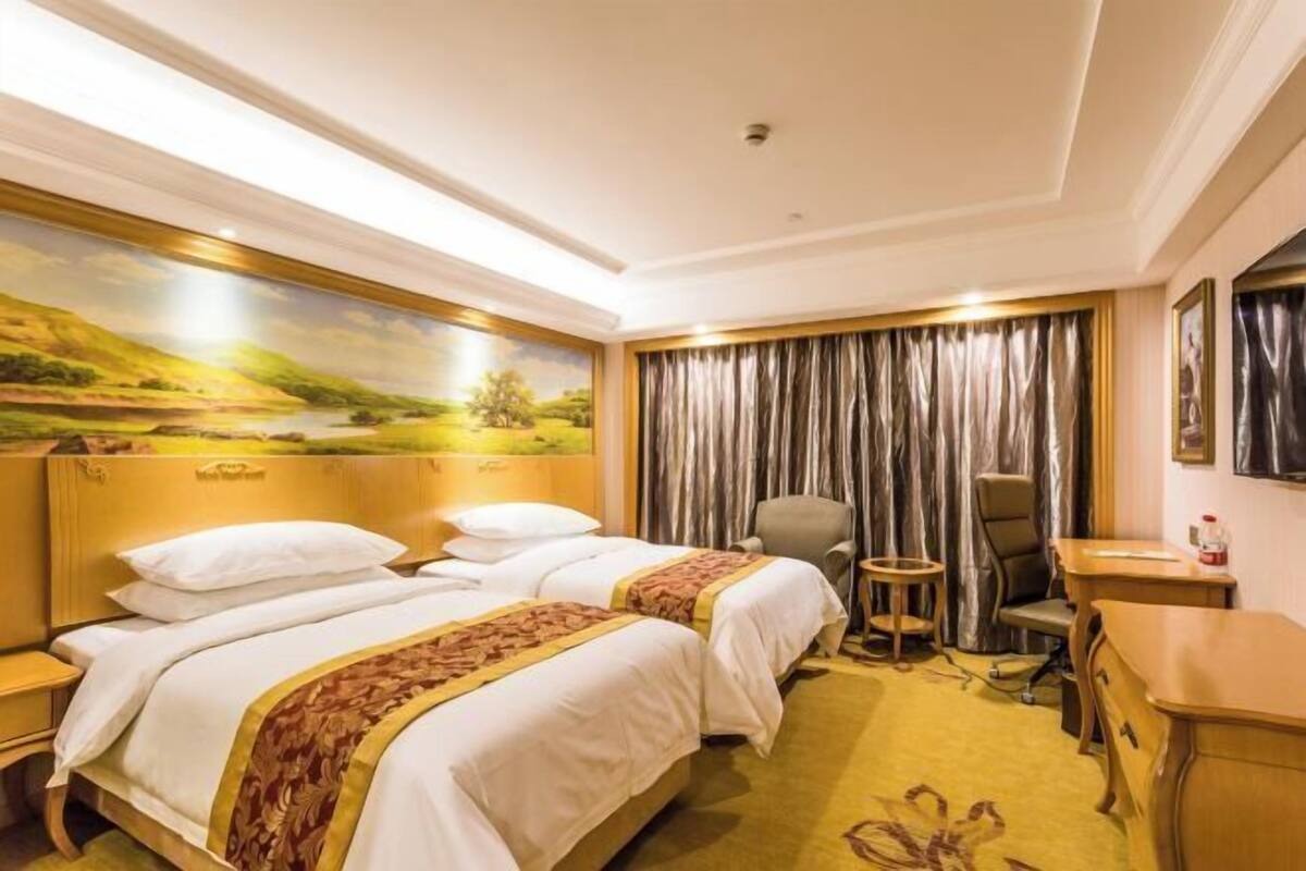 A hotel room with two beds, a desk, a chair, a small table, and a painting on the wall.