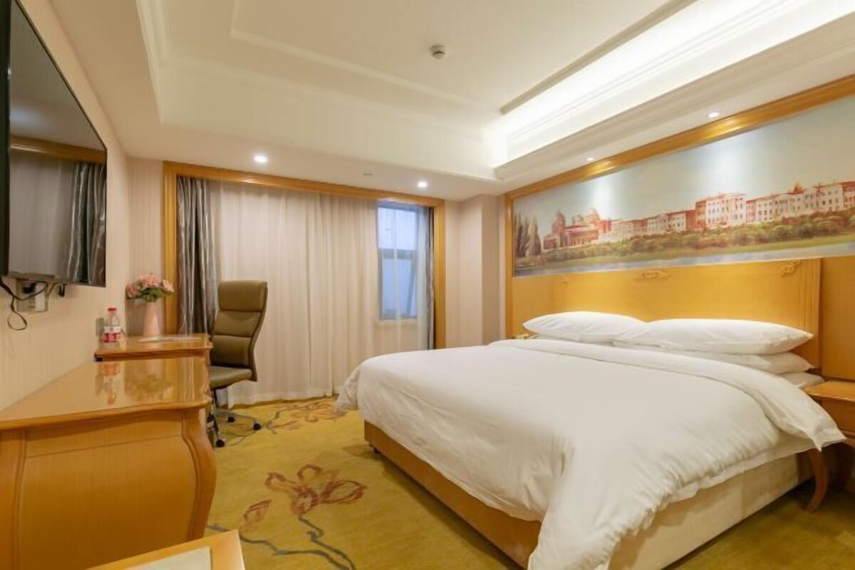 A hotel room with a large bed, a desk, a chair, a painting on the wall, and a window with curtains.