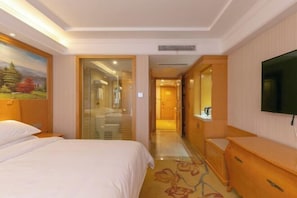 A hotel room with a bed, a TV, a wooden dresser, a painting on the wall, and a hallway leading to another room. - Vienna International Hotel (Shanghai)