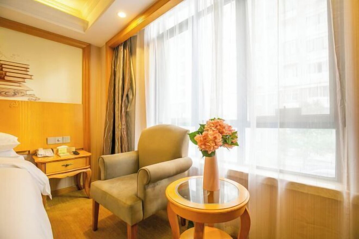 A hotel room with a bed, a chair, a small table with a vase, a nightstand, and a window with curtains.