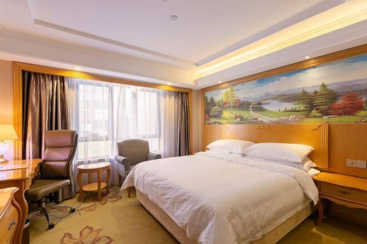 A hotel room with a large bed, a desk with a chair, a small table, and a painting of a landscape on the wall.