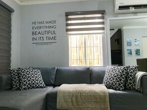 A living room with a grey sofa, patterned pillows, a quote on the wall, and a window with blinds.
