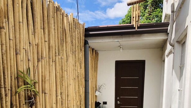 Eine kleine Terrasse im Freien mit einem runden Tisch und weißen Stühlen, einem Bambuszaun, Topfpflanzen und einer braunen Tür.