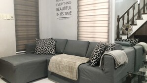 Ein modernes Wohnzimmer mit einem grauen Ecksofa, gemusterten Zierkissen und einer weißen Decke.