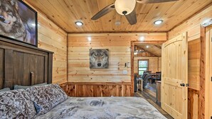 A wooden cabin interior with a bed, a ceiling fan, a mounted TV, and a wooden door. - Cozy Broken Bow Cabin, Walk to the Lukfata Creek! (Broken Bow)