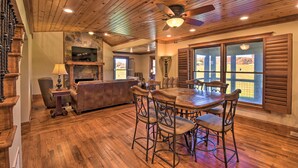 A spacious living room with a wooden ceiling, a fireplace, a dining area with a table and chairs, and large windows with shutters.