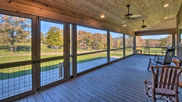 A covered outdoor deck with wooden flooring, large windows, and a view of a green landscape.