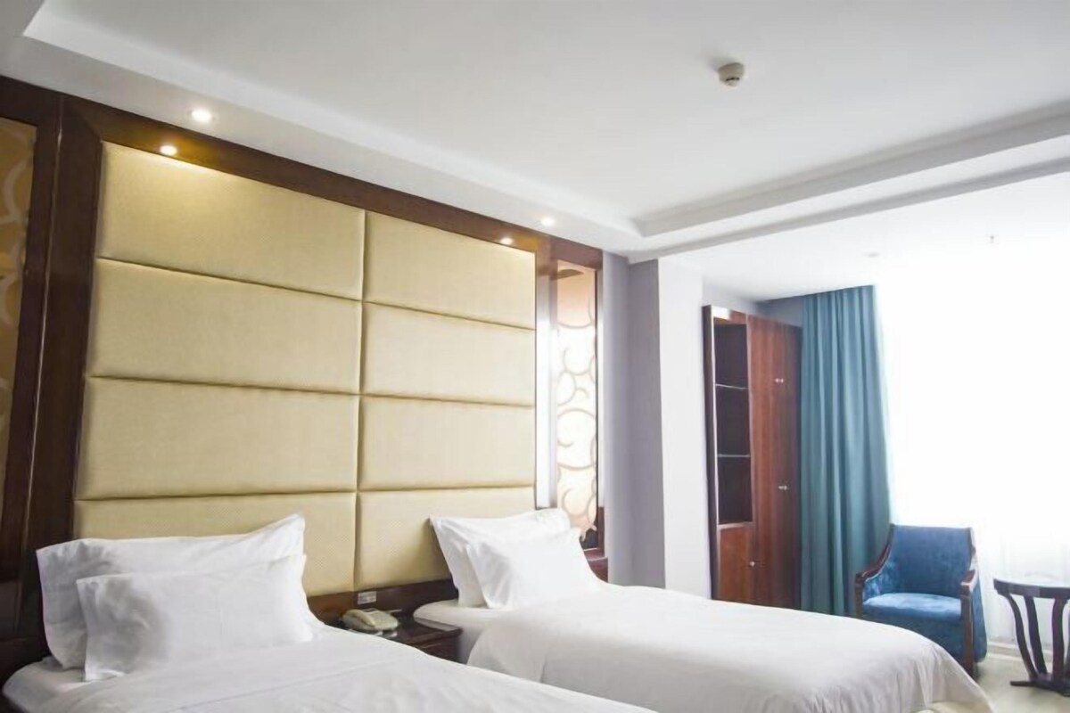A hotel room with two beds, a large padded headboard, a wooden wardrobe, a blue chair, and a small table.