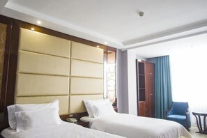 A hotel room with two beds, a large padded headboard, a wooden wardrobe, a blue chair, and a small table. - Lingzhou International Hotel (Fang County)