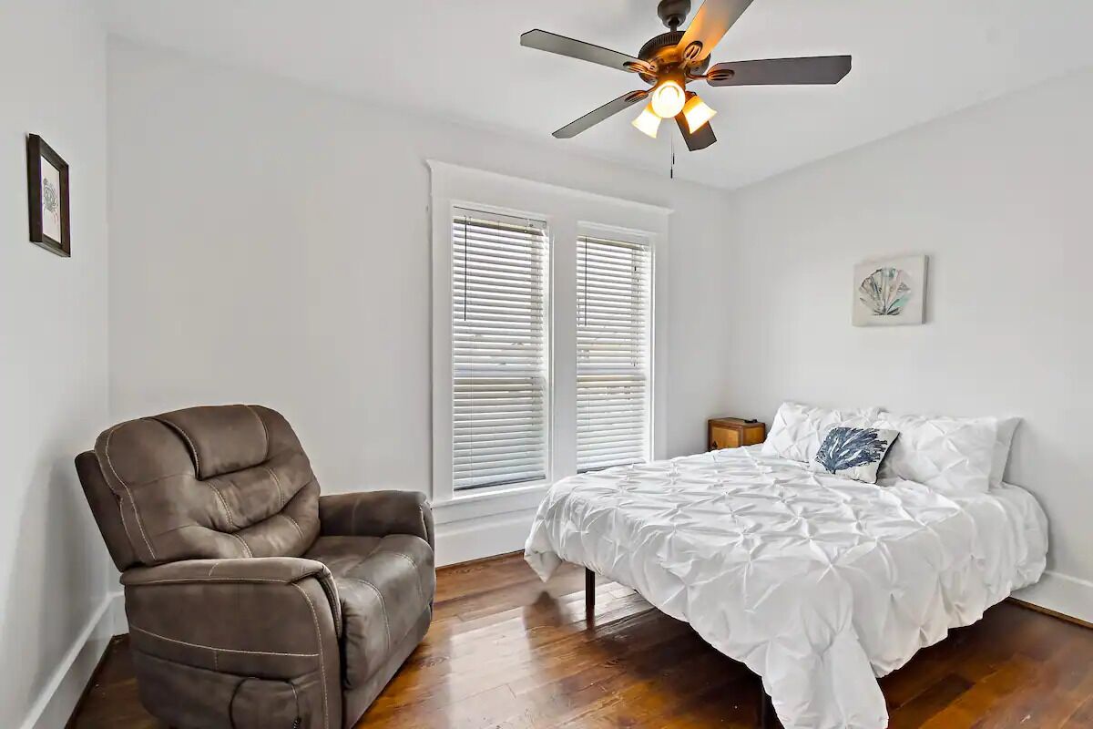 A bedroom with a bed, a recliner, a ceiling fan, and a window with blinds.
