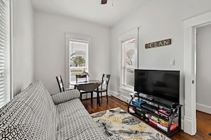 Un salon avec un canapé à motifs, un téléviseur posé sur un meuble, une table à manger avec des chaises et une fenêtre avec des stores.