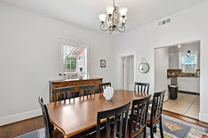 Une salle à manger avec une table et des chaises en bois, un lustre et une vue sur la cuisine par une porte ouverte.