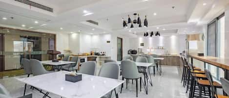 Un comedor moderno con mesas y sillas, una cocina al fondo e iluminación colgante.