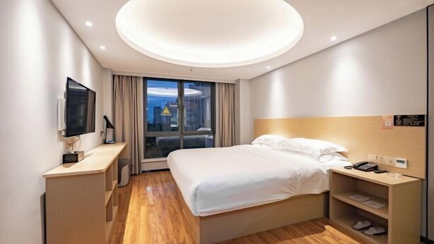 Een moderne hotelkamer met een groot bed, een bureau met een flatscreen-televisie, een nachtkastje met een telefoon en uitzicht op de stad door grote ramen.