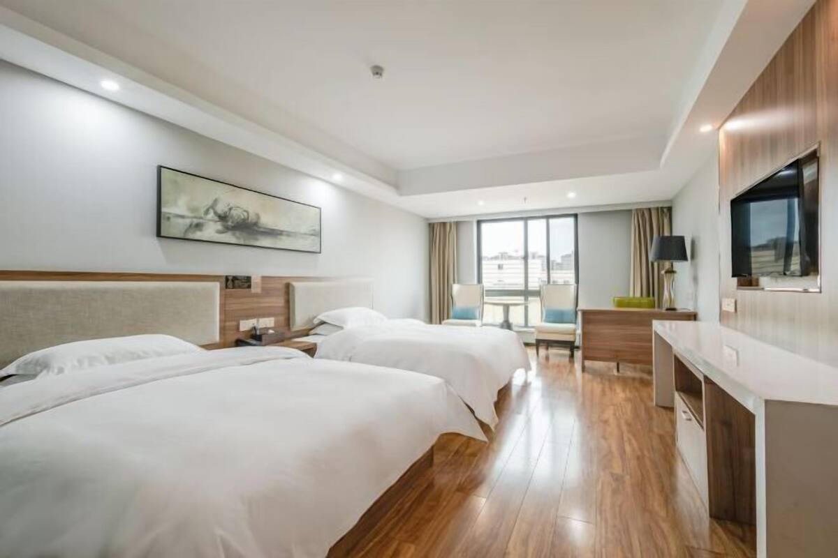 Habitación de hotel con dos camas, un escritorio, una silla y un amplio ventanal con vistas a la ciudad.